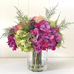 Assorted hydrangea arrangement in a cylindrical glass vase with ferns and sedum.